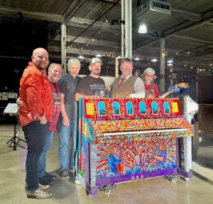 Six men stand behind painted piano
