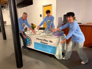 Three men in matching T-Shirts gesture to a public piano sign